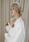 A blonde woman in a white suit poses against a white curtain wearing colorful Wylder boutonnieres on her wrist and lapel