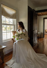 A bride holds a Haven bridal bouquet and poses while looking out a window