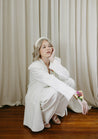 A woman in a white suit poses against a white curtain, wearing a Wylder corsage