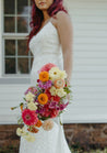 A bride with red hair stands just out of focus, holding a colorful Wylder bridal bouquet