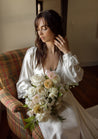 A brunette woman sits in a plaid chair, holding a cream and white bridal bouquet; Haven floral collection